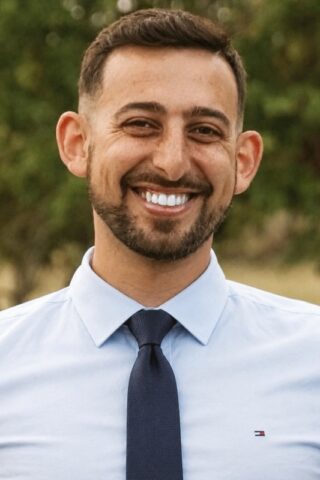 Dr Rami 570x800 - Chiropractor in San Mateo | Neurolink Chiropractic | Accepting New Patients Smiling man with short dark hair and a beard, wearing a light blue dress shirt and navy tie, outdoors with greenery in the background.
