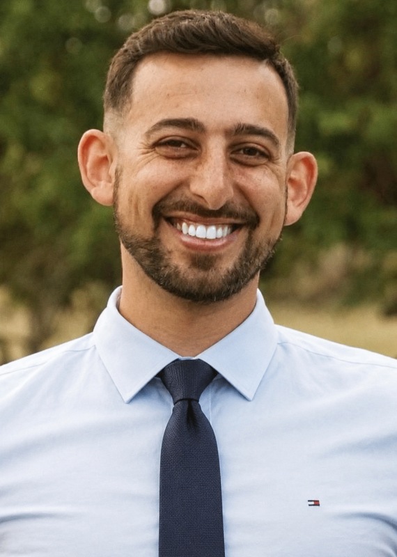 Dr Rami 570x800 - Chiropractor in San Mateo | Neurolink Chiropractic | Accepting New Patients Smiling man with short dark hair and a beard, wearing a light blue dress shirt and navy tie, outdoors with greenery in the background.
